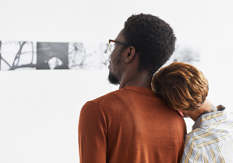 Graphic portrait of mixed-race couple embracing while looking at paintings at modern art gallery exhibition, copy space