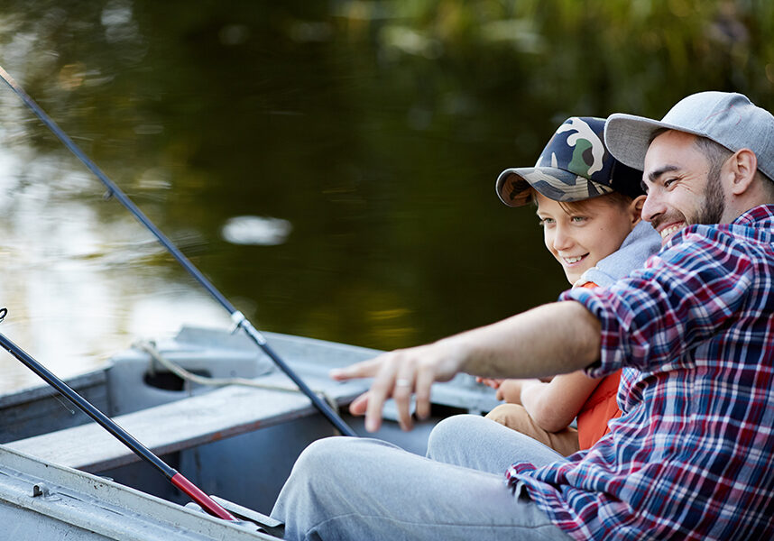 Cheerful father showing to his son a good place for fishing while they sitting in the boat